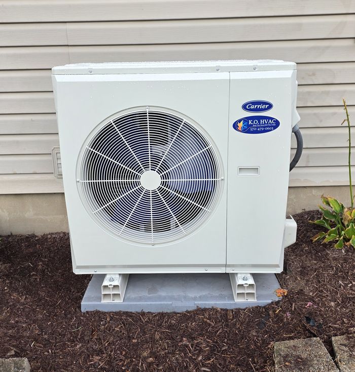 A white Carrier outdoor unit with a KO HVAC label, on a gray pad in a mulch bed against beige siding