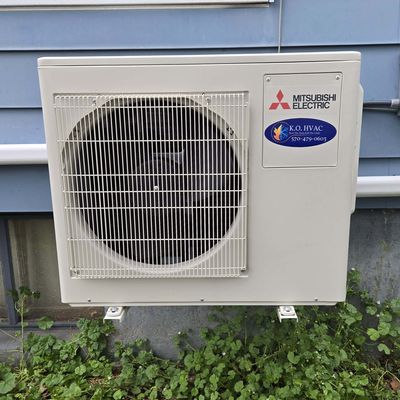 A Mitsubishi Electric outdoor unit with a KO HVAC label, mounted on wall brackets against blue siding