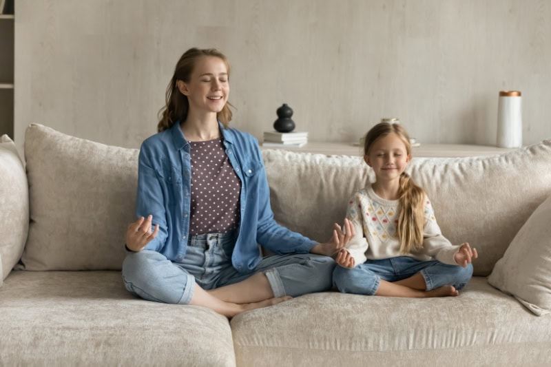 Air Conditioning Noise Concerns. A photo of a relaxed mother and young daughter sitting in lotus position on cozy couch with closed eyes.
