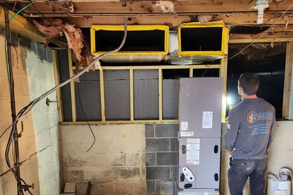 A KO HVAC technician beside a new air handler in a stripped basement, new duct openings framed above