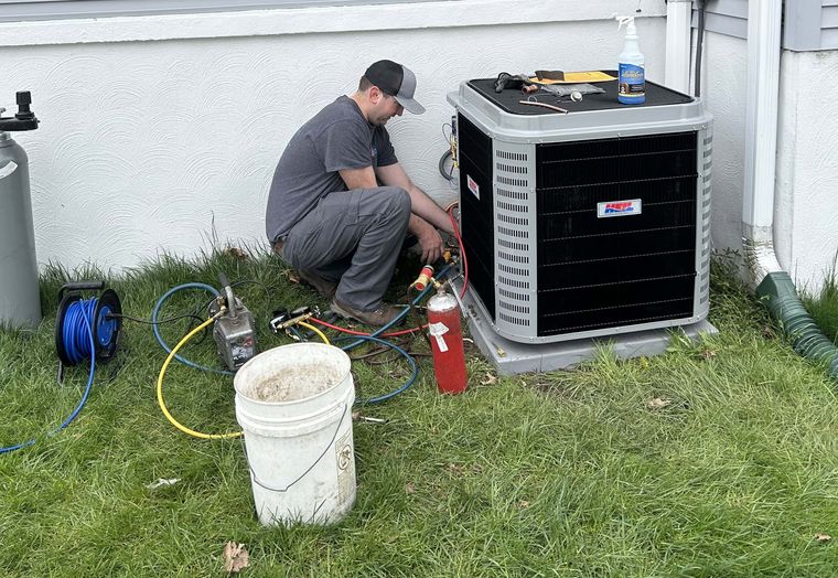 A KO HVAC technician crouched beside a condenser with gauges and hoses on a lawn