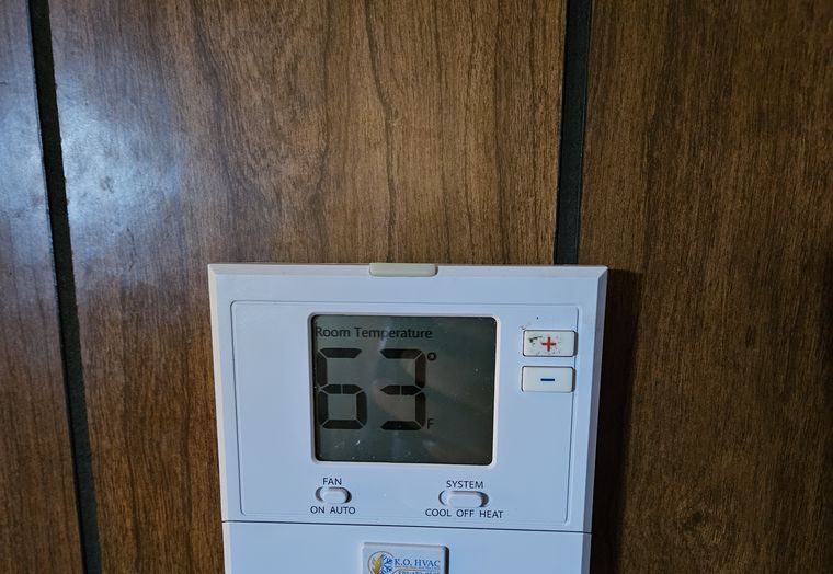 A wall thermostat on wood paneling reading 63, with a KO HVAC sticker