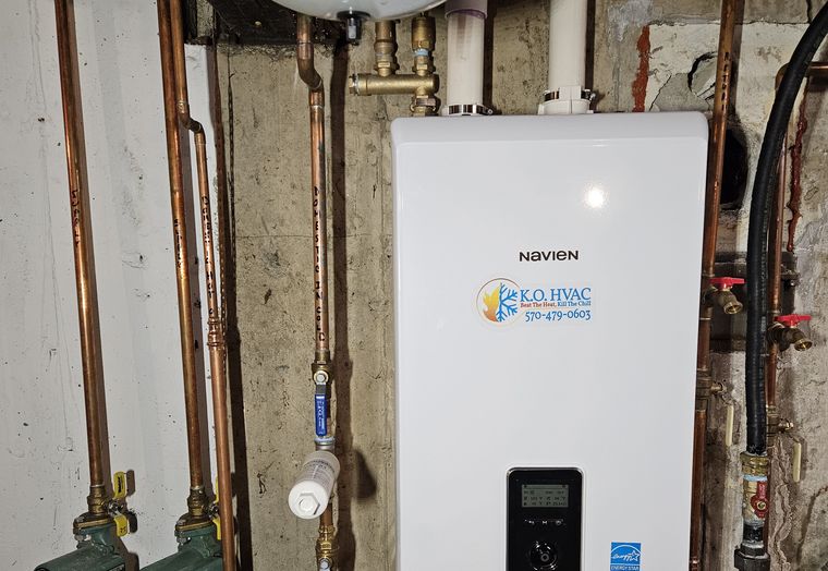 A Navien tankless water heater with a KO HVAC sticker on a basement wall, with copper piping