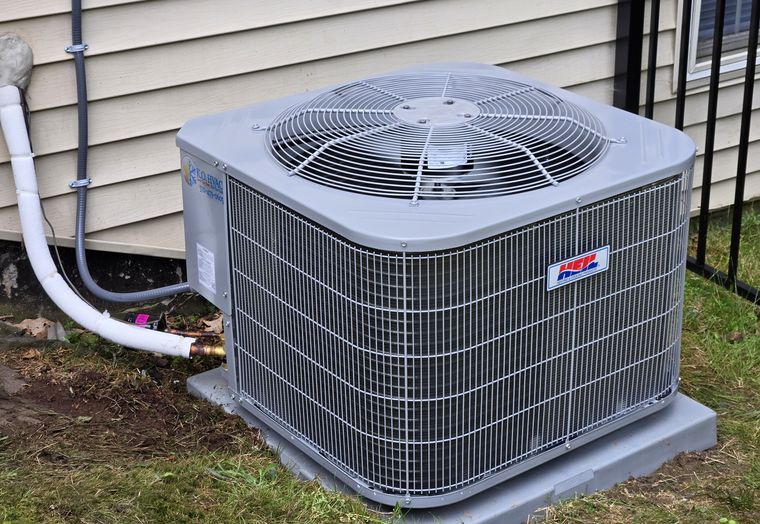 A new condenser on a pad beside a house, its refrigerant line in a white cover