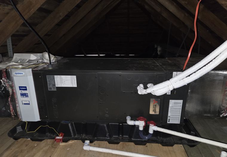 A horizontal air handler on a drain pan in an attic, with white line-set covers