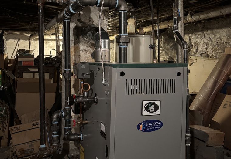 A gray boiler with black iron piping and a KO HVAC sticker in an old basement