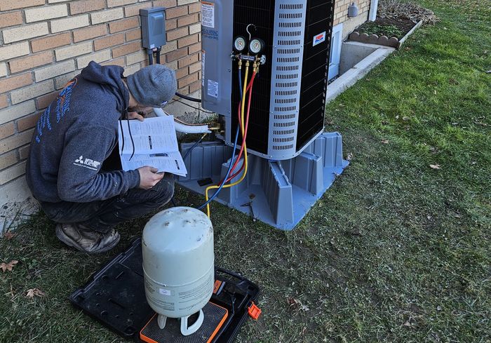 A KO HVAC technician crouched at an outdoor unit with gauges and a recovery tank