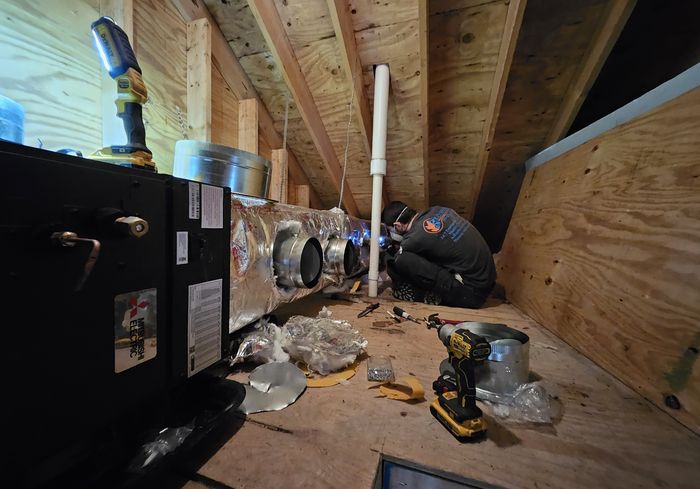A KO HVAC technician installing a black air handler and duct collars in an attic