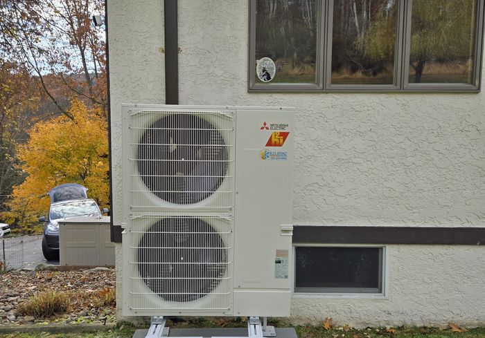 A two-fan Mitsubishi outdoor unit on a stand beside a stucco house in autumn