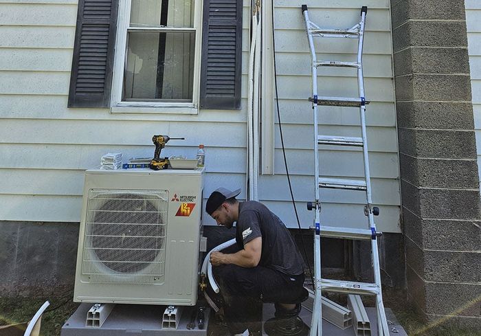 A KO HVAC technician crouched at a mini-split outdoor unit under a ladder