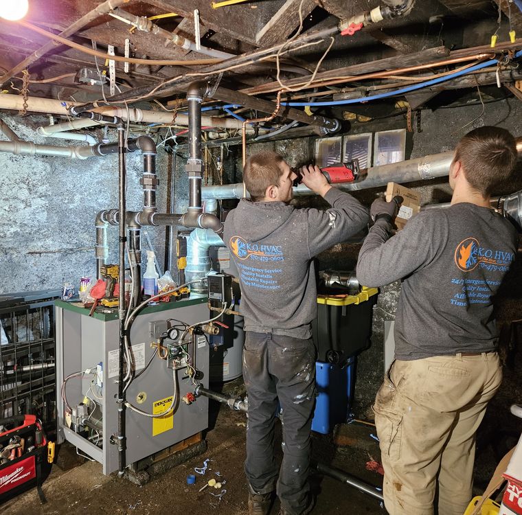 Two KO HVAC technicians in company shirts working on a heating system in a basement