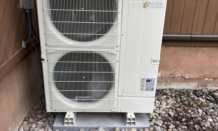 A two-fan Mitsubishi outdoor unit on a rock bed against wood siding